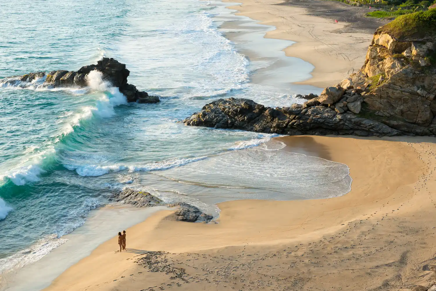 Two people walk along a sandy beach near rocky cliffs