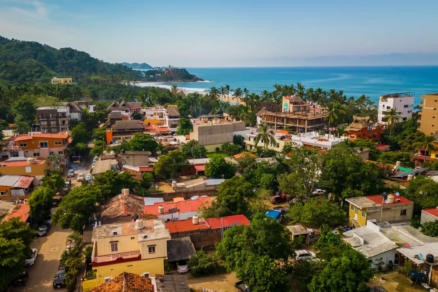 A scenic aerial view of a coastal village with buildings surrounded by lush green trees hills in the background and the ocean nearby