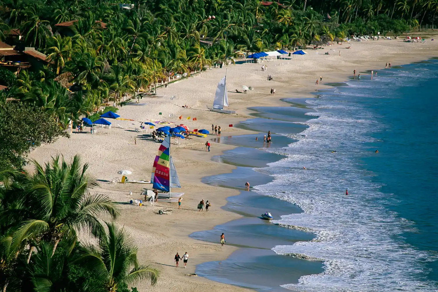 A beach with people umbrellas and a sailboat visible